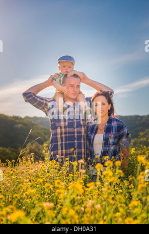 A young Caucasian family, father, mother and toddler boy on daddy's shoulders, walking through a field of yellow wildflowers. Stock Photo