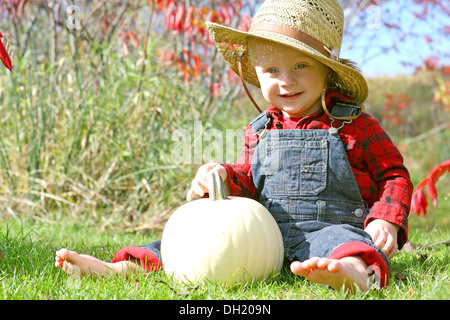 a cute, smiling baby boy is sitting outside in the grass with a white pumpkin on a sunny autumn day, dressed like a farmer Stock Photo