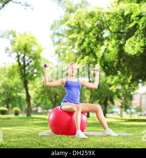 Blond female athlete in a park sitting on a pilates ball and exercising with dumbbells Stock Photo