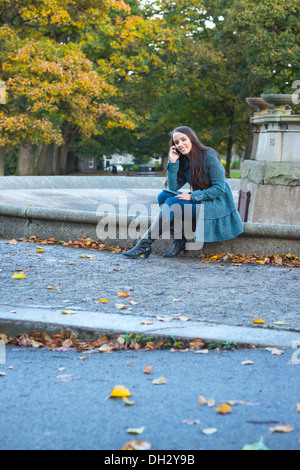 Young woman in a park during autumn sitting on the edge of a fountain using an apple iphone to make a call Stock Photo
