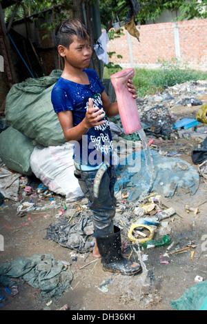 A young boy is pouring water out of his pink rubber boot while working at a garbage recycling depot  in Phnom Penh, Cambodia. Stock Photo