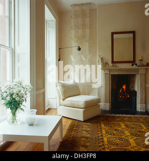 White chair and coffee table in white living room with yellow oriental rug and Georgian fireplace with lit fire Stock Photo