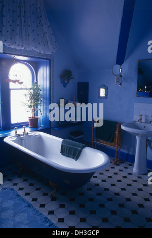 Roll top bath below window in bright blue bathroom with black+white tiled floor and white pedestal basin Stock Photo