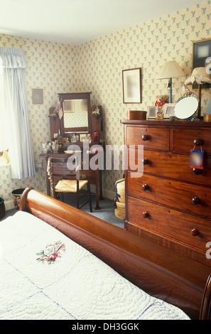 Victorian mahogany chest-of-drawers and sleigh bed in cottage bedroom with patterned wallpaper and antique dressing table Stock Photo