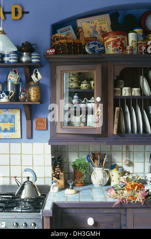 Dark wood fitted unit with glass cupboard door above tiled worktop beside Alessi kettle on range oven in cluttered blue kitchen Stock Photo