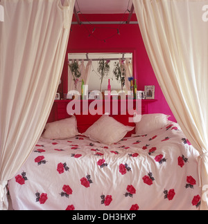White drapes on simple four poster bed with white cushions and rose-patterned duvet below mirror on shelf in pink bedroom Stock Photo