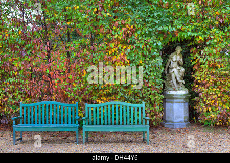 Two wooden benches on alley in front of bushes with lush colorful foliage and old sculpture in autumn Racconigi park, Italy. Stock Photo