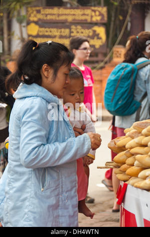 Vertical portrait of a local Lao lady and her baby buying baguettes at a street market in Laos. Stock Photo