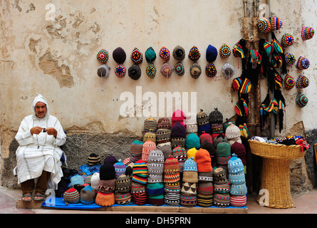 Street vendor wearing a traditional Djellabah while knitting a cap, next to him are his hats out for sale, Essaouira, Morocco Stock Photo