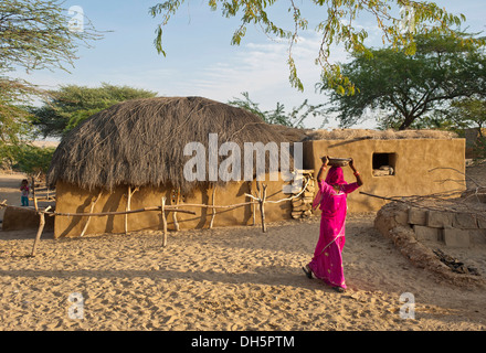 An Indian woman wearing a traditional sari and a veil over her face is carrying a bowl on her head, village in the Thar Desert Stock Photo