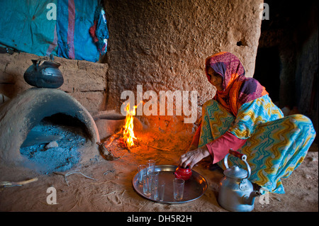 Elderly Berber woman preparing tea while squatting on the ground in front of a clay oven and a small fire, Erg Chebbi Stock Photo