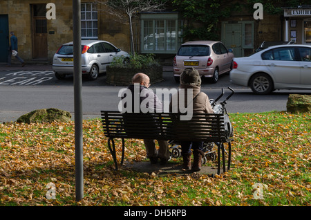 An elderly couple sitting on a bench with pushchair surrounded by fallen autumn leaves Stock Photo