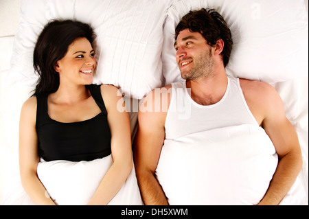 Beautiful couple laid in a white bed next to each other smiling Stock Photo