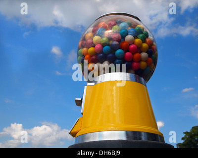 A vintage yellow gumball machine with multi-colored gumballs stands tall in front of a blue sky with wispy clouds. Stock Photo