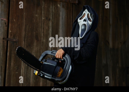 Man in scary 'scream' mask holds chainsaw. Mask of grim reaper. Carnival white ghost mask and black hood. Scary Movie film. Stock Photo