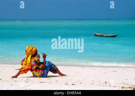 Woman in traditional dress sitting on the beach looking at the sea, Zanzibar, Tanzania Stock Photo