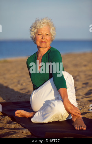 Senior woman enjoying yoga on the beach at morning. Elderly woman doing stretching exercises on beach. Stock Photo