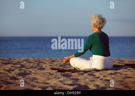 Side view of elderly woman in meditation on the beach. Senior lady sitting on the beach in lotus pose doing relaxation exercise. Stock Photo