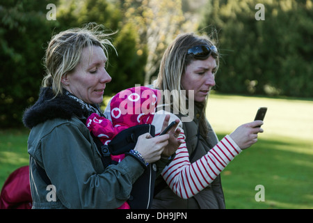 Two young mothers outside on a sunny day using smart mobile phones Stock Photo