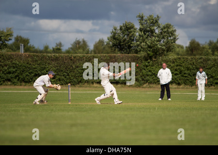 Village cricket. The batsman pulls the ball square of the wicket with the wicketkeeper waiting to pounce whilst a fielder and an Stock Photo