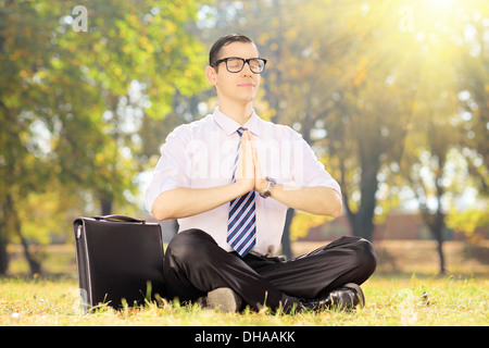 Young businessperson with tie doing yoga exercise seated on a grass in a park Stock Photo