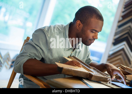 A young man at his workbench in a picture framing studio. Surrounded by samples. Using a digital tablet. Stock Photo