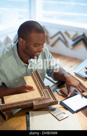 A young man at his workbench in a picture framing studio. Surrounded by samples. Using a digital tablet. Stock Photo