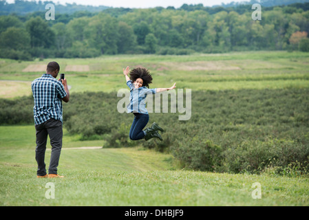 A man taking a photograph of a woman leaping in the air, jumping for joy with her arms outstretched. Stock Photo