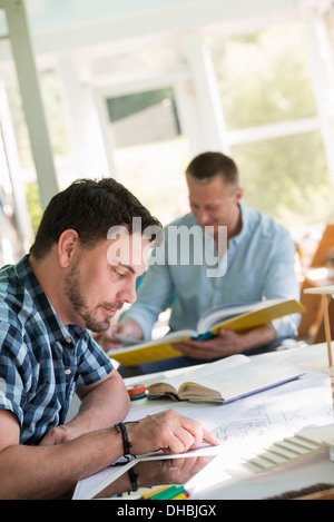 Two men seated at a table using books and a digital tablet. Stock Photo