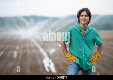 An organic vegetable farm, with water sprinklers irrigating the fields. A man in working clothes with his hands on his hips. Stock Photo