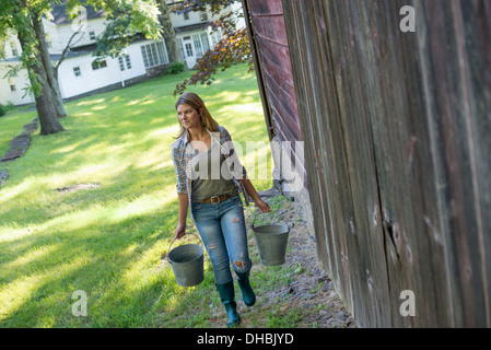 A woman carrying a bucket for animal feed in each hand, outside a barn. Stock Photo