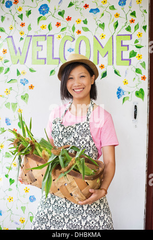 Working on an organic farm. A young Asian woman by the Welcome sign with a large basket of vegetables, freshly picked. Stock Photo