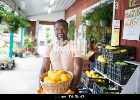 Working on an organic farm. A man carrying a large basket of yellow squash vegetables. Displays of fresh produce for sale. Stock Photo