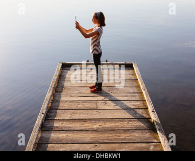 A young girl holding a digital tablet in front of her, standing on a wooden dock over the water. Stock Photo