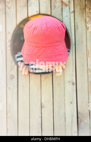 Childhood lifestyle moment. Little girl in read hat looking down from round window in tree house Stock Photo