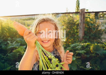 A young girl with long red curly hair outdoors in a garden holding a large fresh picked carrot.