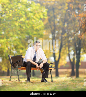 Disappointed young businessman sitting on a wooden bench with bottle in his hand, in park Stock Photo