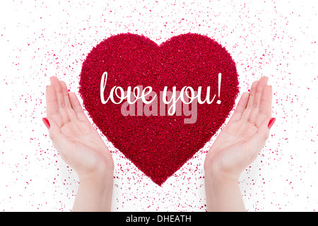 On the red heart made of sand written 'love you'. Nearby there are women's hands with red manicure. Stock Photo