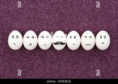 Seven eggs with different facial expressions are lying on the decorative purple sand. Stock Photo