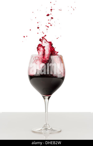 Splash of red wine in glass. Nearby there are splatter of wine. A glass stands on a glossy gray surface and isolated on white Stock Photo