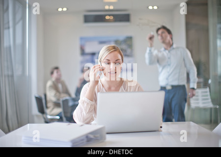 Woman sitting at conference room table using laptop and mobile phone, colleagues working in background Stock Photo
