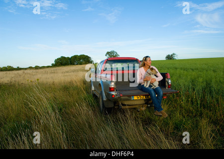 Woman sitting on back of pick up truck in a field, holding a dog Stock Photo