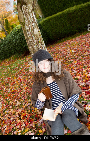 Vertical photo of teenage girl, looking forward, enjoying her book, and drink, while lying against a tree during the autumn Stock Photo
