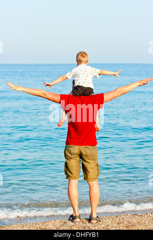 Son sits on his father's shoulders his arms to the side against the sea Stock Photo