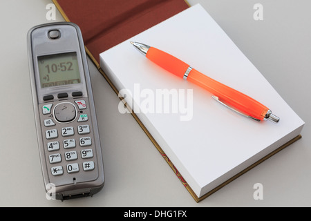 Cordless digital telephone phone handset with a blank notepad notebook page and an orange pen on a plain grey desktop background Stock Photo
