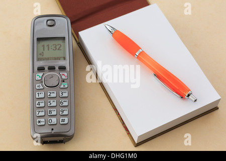 Cordless modern digital telephone handset with a blank notepad notebook and a pen on a plain beige tabletop background Stock Photo