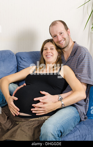 Smiling man embracing happy pregnant woman sitting on couch holding tummy Stock Photo