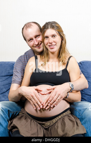 Happy parents make heart sign with two pairs of hands on pregnant woman's belly sitting on couch Stock Photo