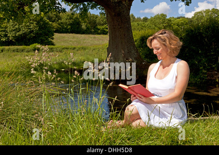 A woman in a white dress sat by a stream reading a book on a bright summers day. Stock Photo