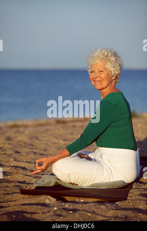 Happy old woman going relaxation exercise while sitting on sandy beach. Senior caucasian practicing yoga by the ocean in morning Stock Photo